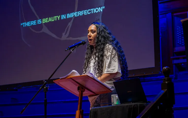 A woman speaking at a lectern on a stage lit blue, under the words There is beauty in imperfection